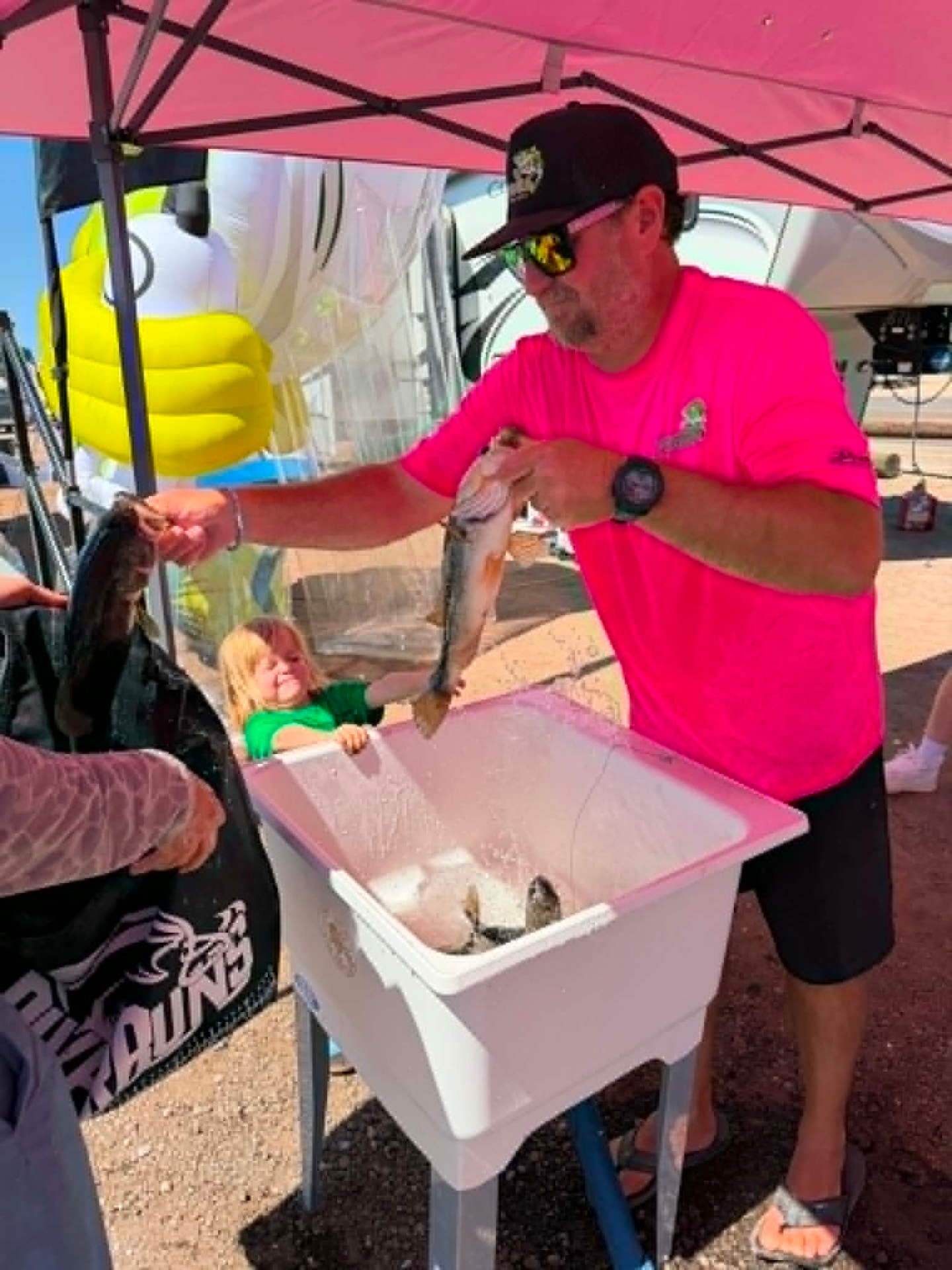 An angler in a pink shirt holding a bass over a tub at the weigh-in while a child looks on